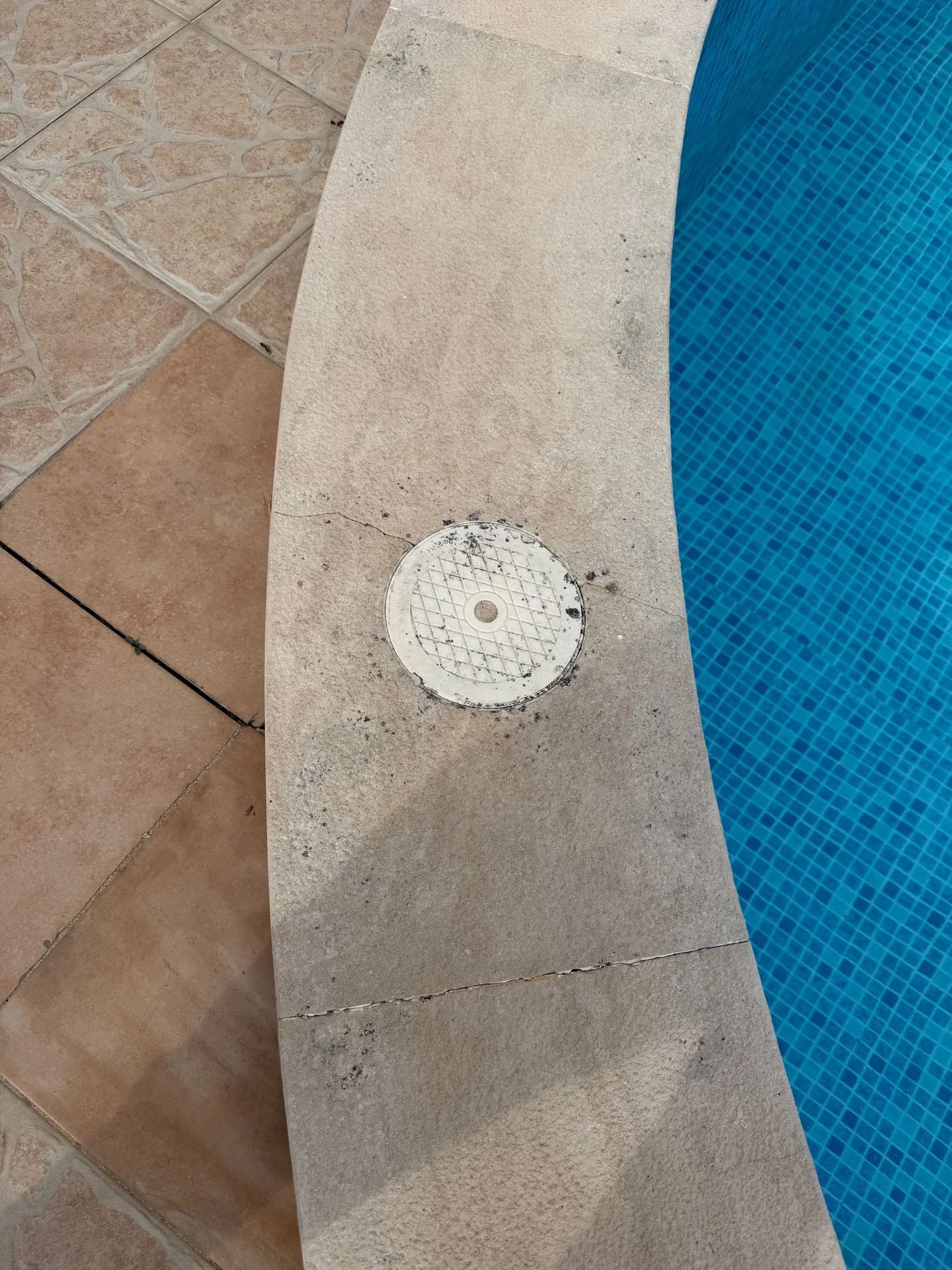 Borda de piscina clara com fissura visível e tampa circular junto a mosaico azul de piscina